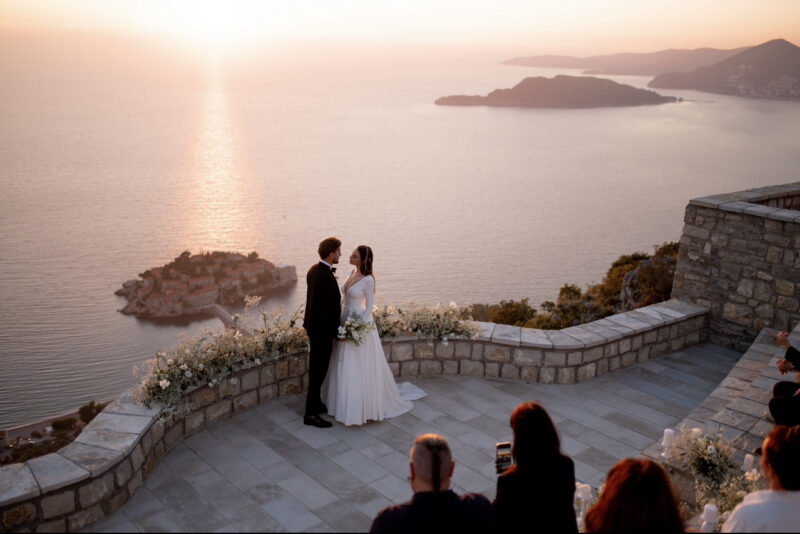 A bride and groom standing on a balcony overlooking the sea.