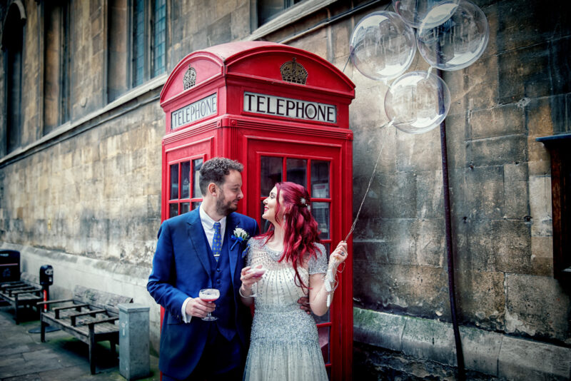 A bride and groom pose in front of a telephone booth.