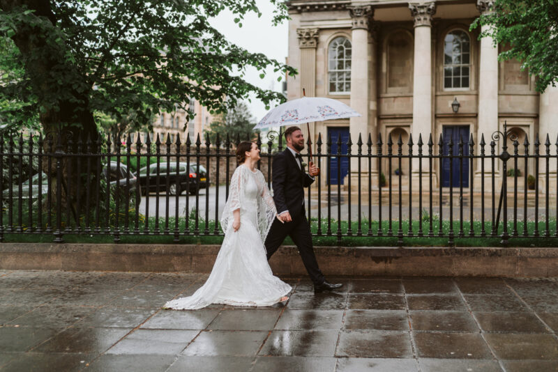 A bride and groom walking down the street with an umbrella.