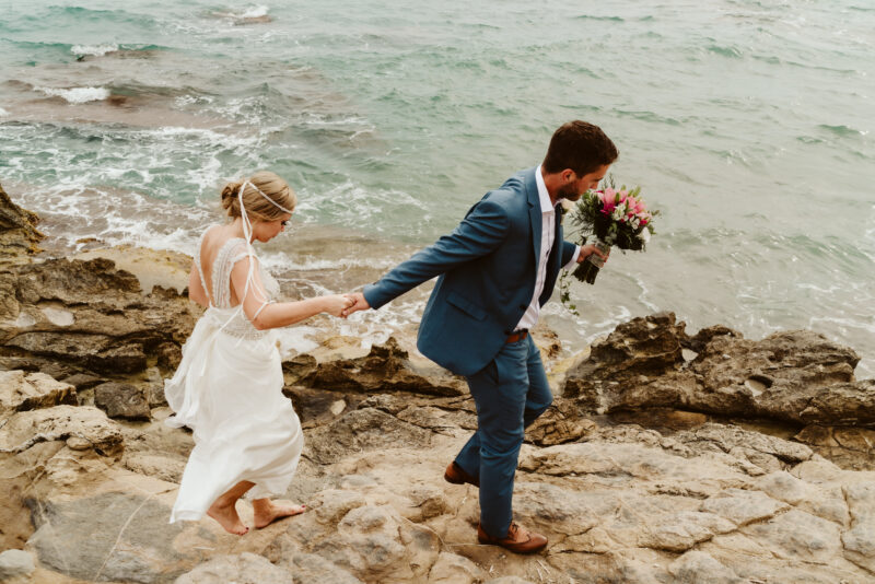 A bride and groom walking down a rocky path near the ocean.