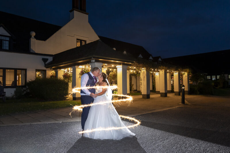 A bride and groom with sparklers in front of a building at night.