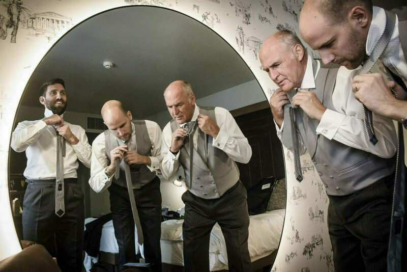 A group of men adjusting their ties in front of a mirror.